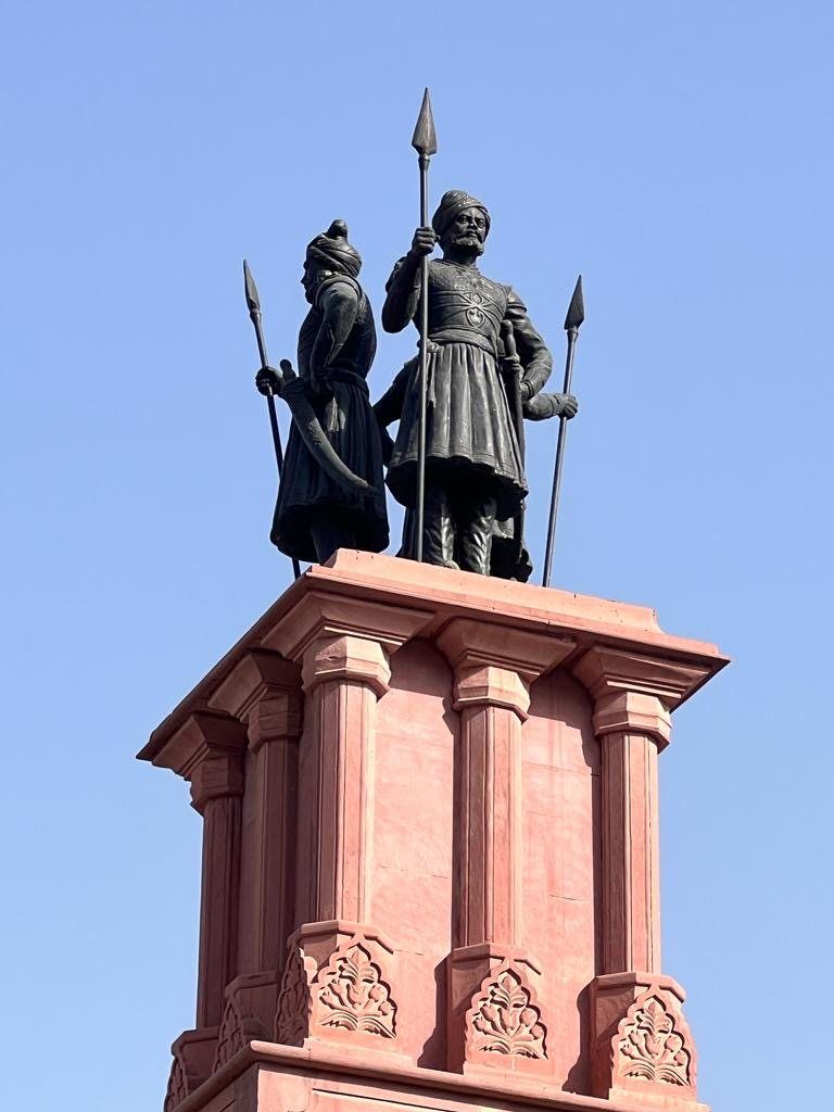 Two dark warrior figures with spears on top of a red sandstone column