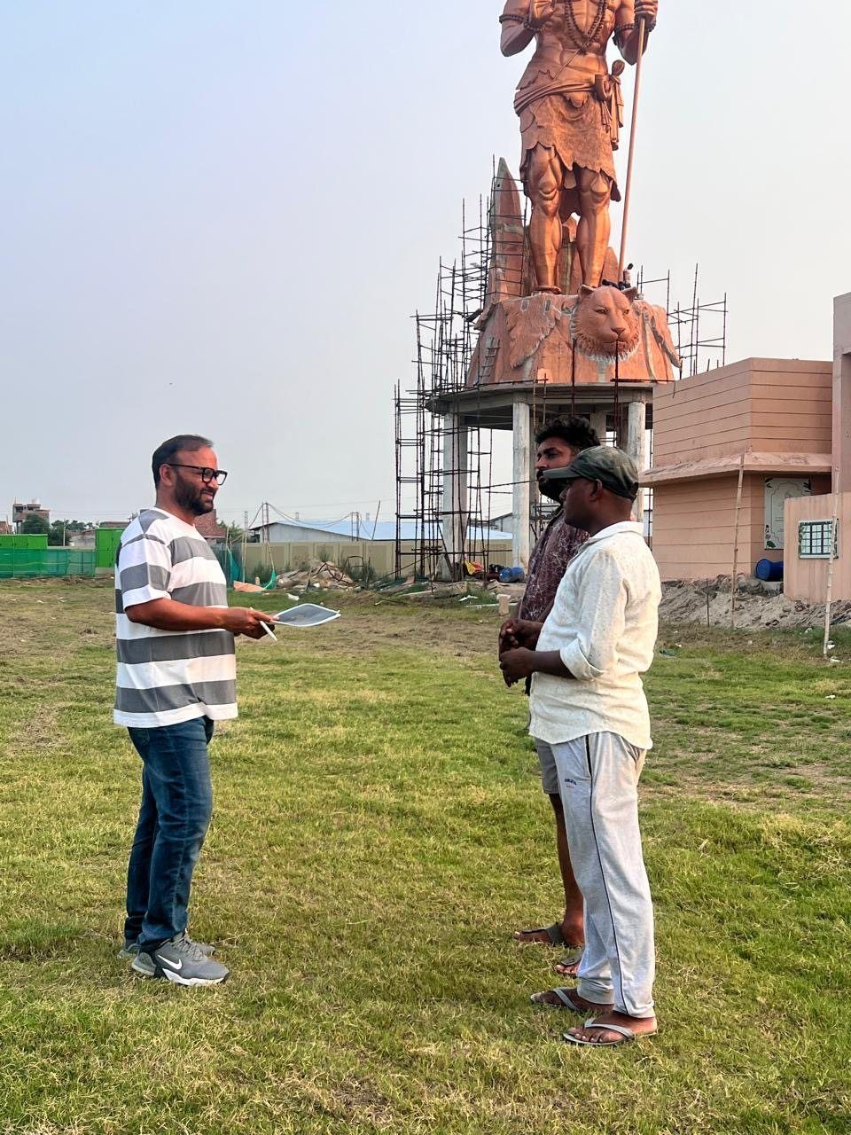 Three men talking in a field below the Shiva sculpture, with scaffolding at its base