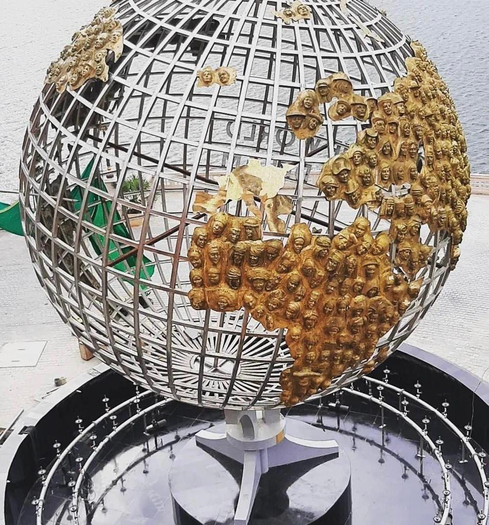 Open steel globe with continents made of sculpted faces, seen from above in a fountain basin