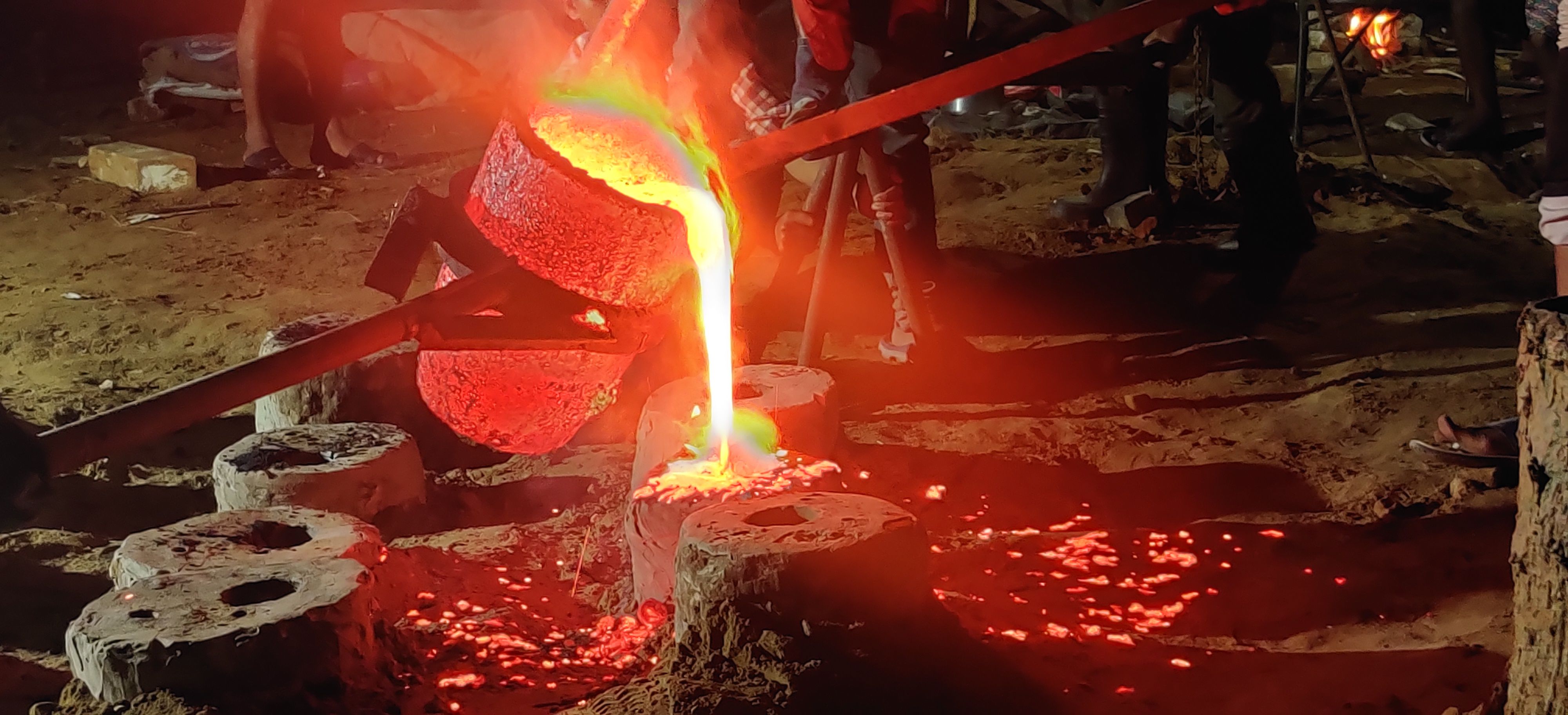 Close view of a crucible pouring a bright stream of molten metal into a mould, sparks on the sand around it
