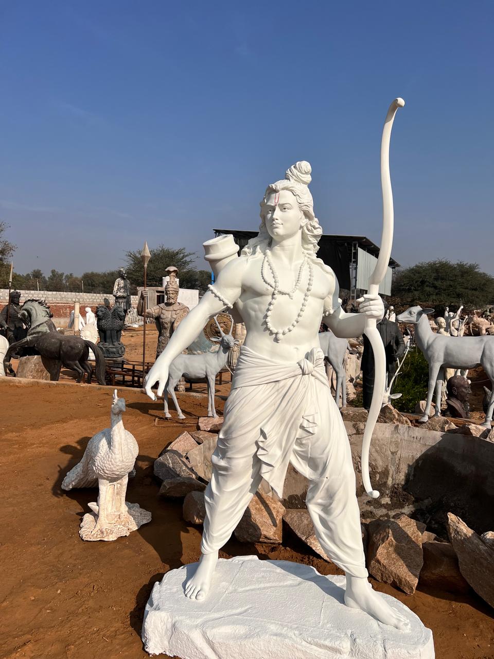 White figure of Lord Ram holding a bow, standing on a rock base among sculptures in the studio yard